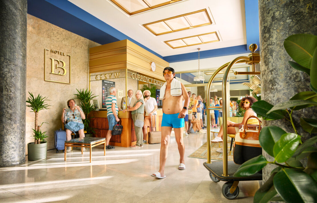 Adult man in swimsuit walks through a busy hotel lobby with a luggage cart; guests line up at the check-in and currency exchange.