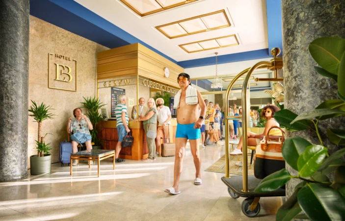 Adult man in swimsuit walks through a busy hotel lobby with a luggage cart; guests line up at the check-in and currency exchange.