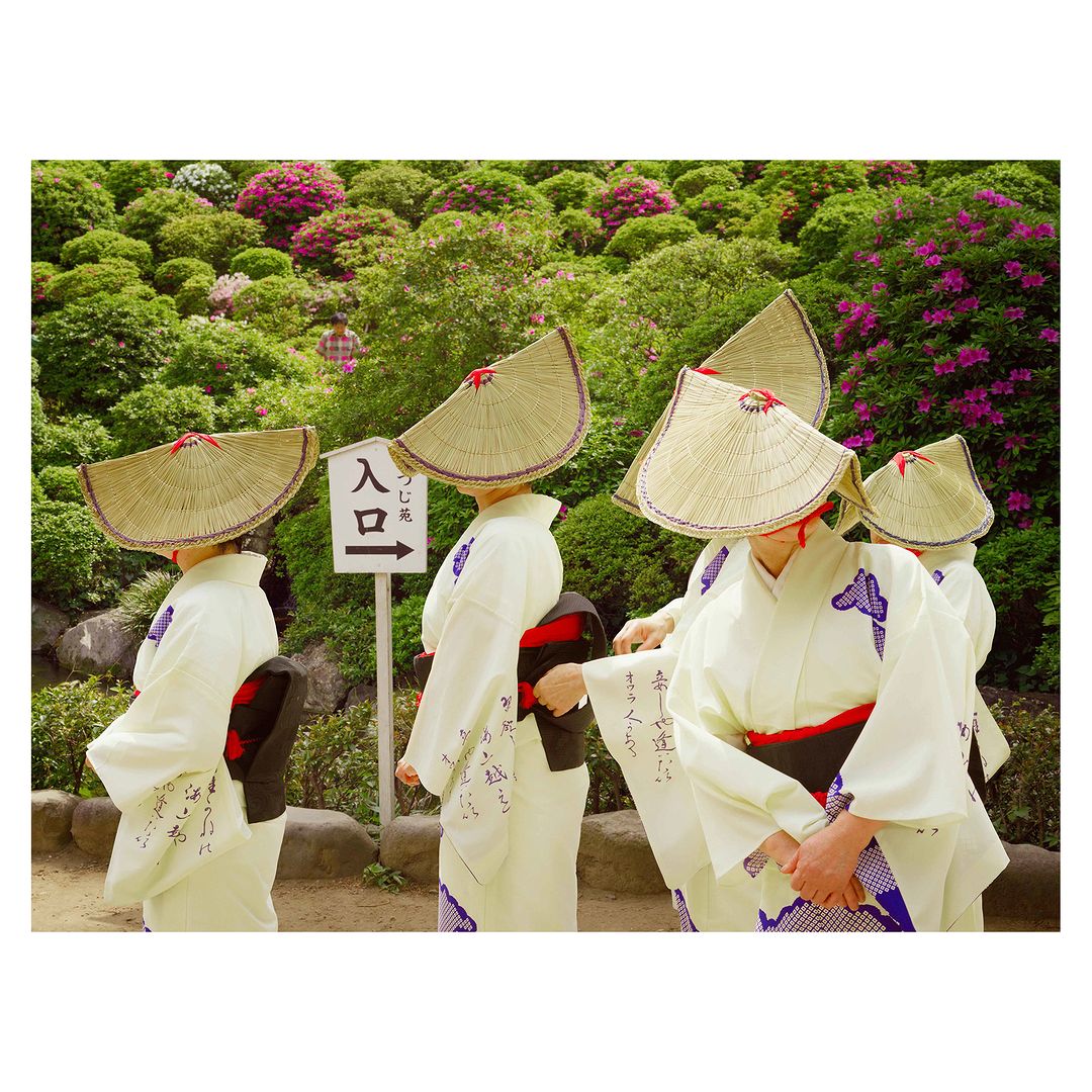 Group of women in kimonos and straw hats standing in a garden with lush greenery and pink flowers, with a sign pointing to an entrance.