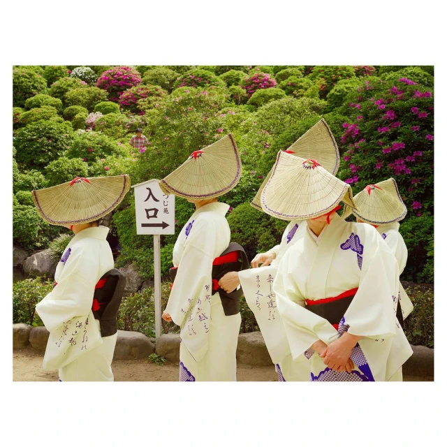 Group of women in kimonos and straw hats standing in a garden with lush greenery and pink flowers, with a sign pointing to an entrance.