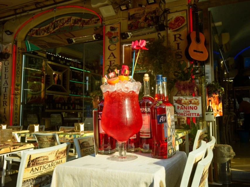 Cocktail drink with orange garnish and crushed ice, sitting on a table in front of a cafe with outdoor seating, bottles, and sign.