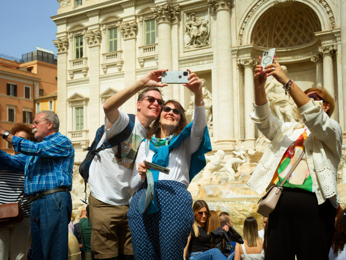 Couple taking a selfie with a smartphone at the Trevi Fountain in Rome, surrounded by a crowd of tourists on a sunny day.