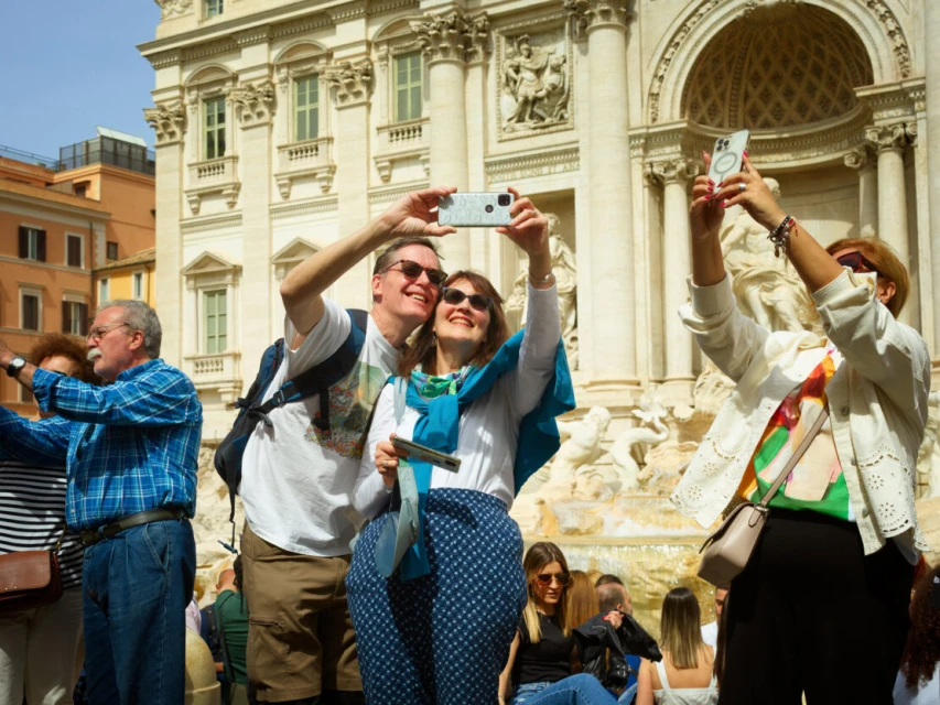 Couple taking a selfie with a smartphone at the Trevi Fountain in Rome, surrounded by a crowd of tourists on a sunny day.