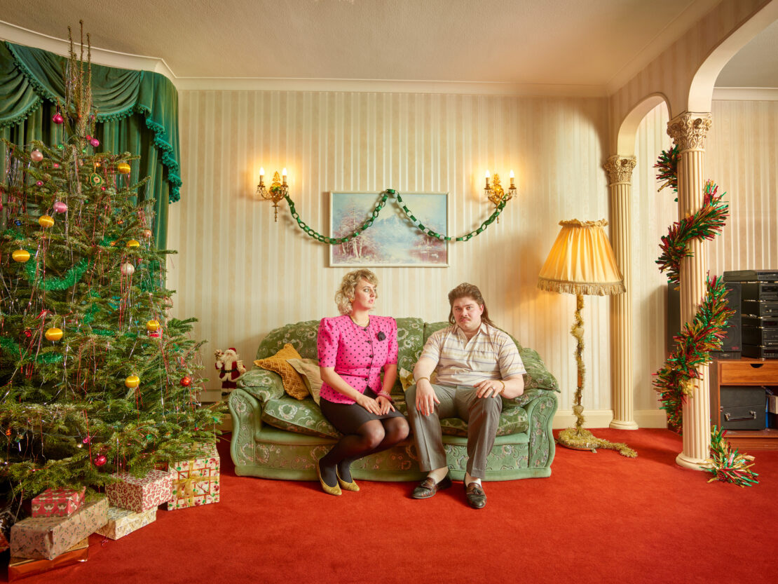 Couple sitting on a green sofa in living room with Christmas tree and retro decor, brightly lit with vintage style.