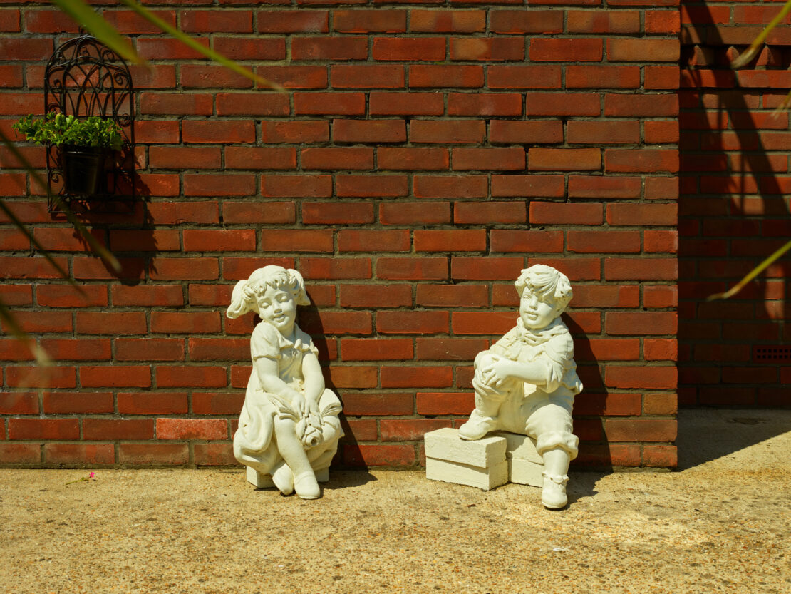 Two concrete statues of children, one boy and one girl, sit beside each other in front of a brick wall under bright sunlight.