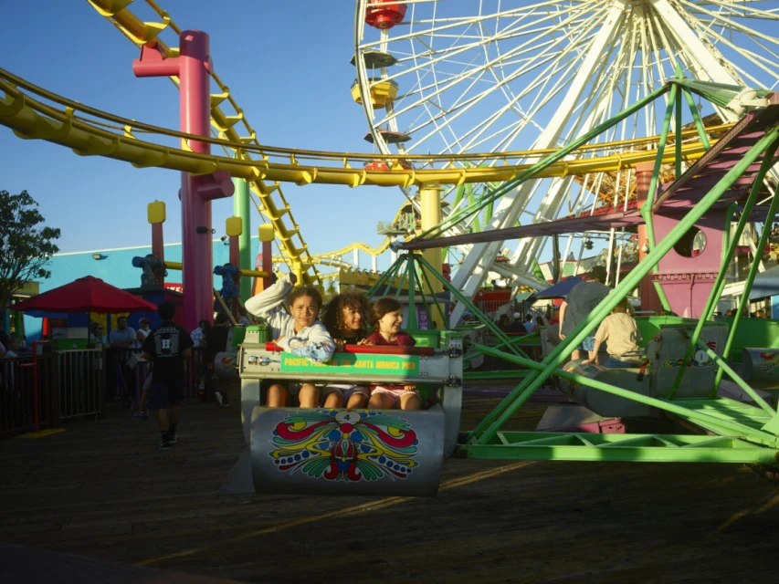 Three young girls ride an amusement park glider, with a Ferris wheel and roller coaster tracks visible against a bright blue sky.