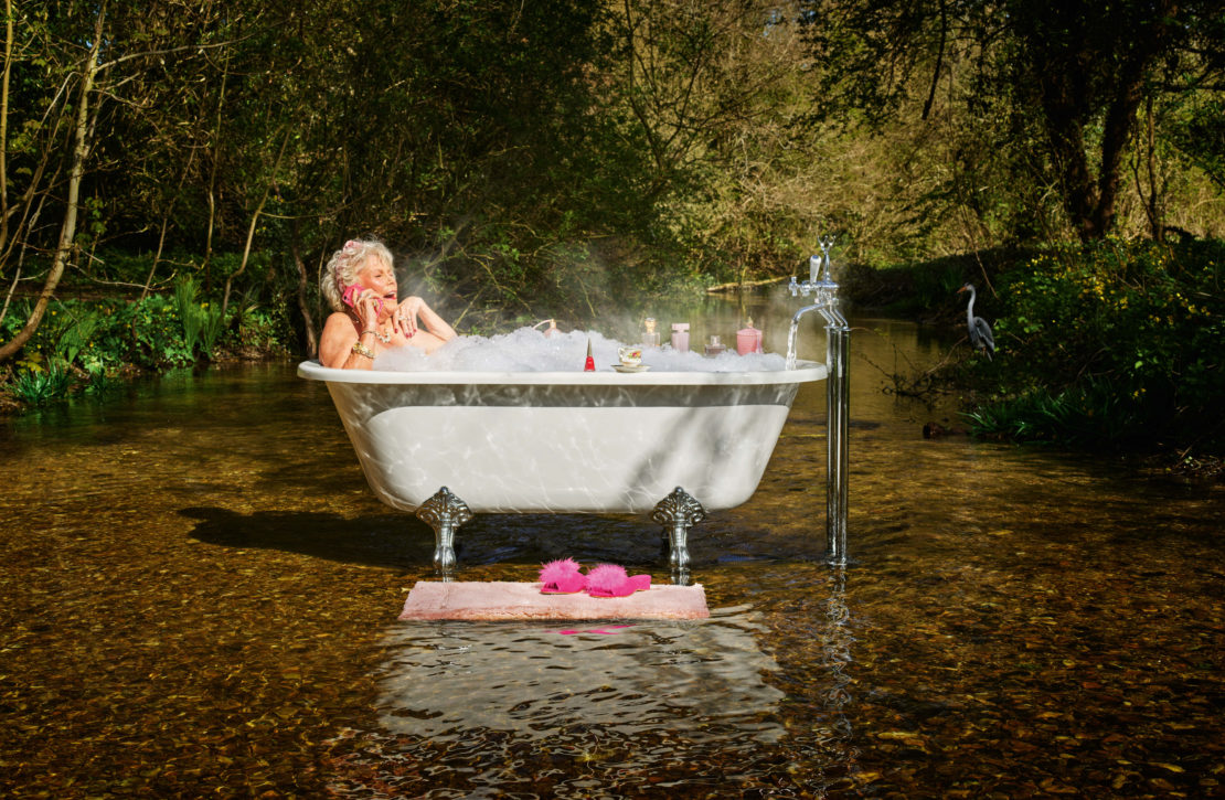 Older woman relaxes in a clawfoot bathtub with foam in a shallow river surrounded by lush trees while talking on a pink cell phone.