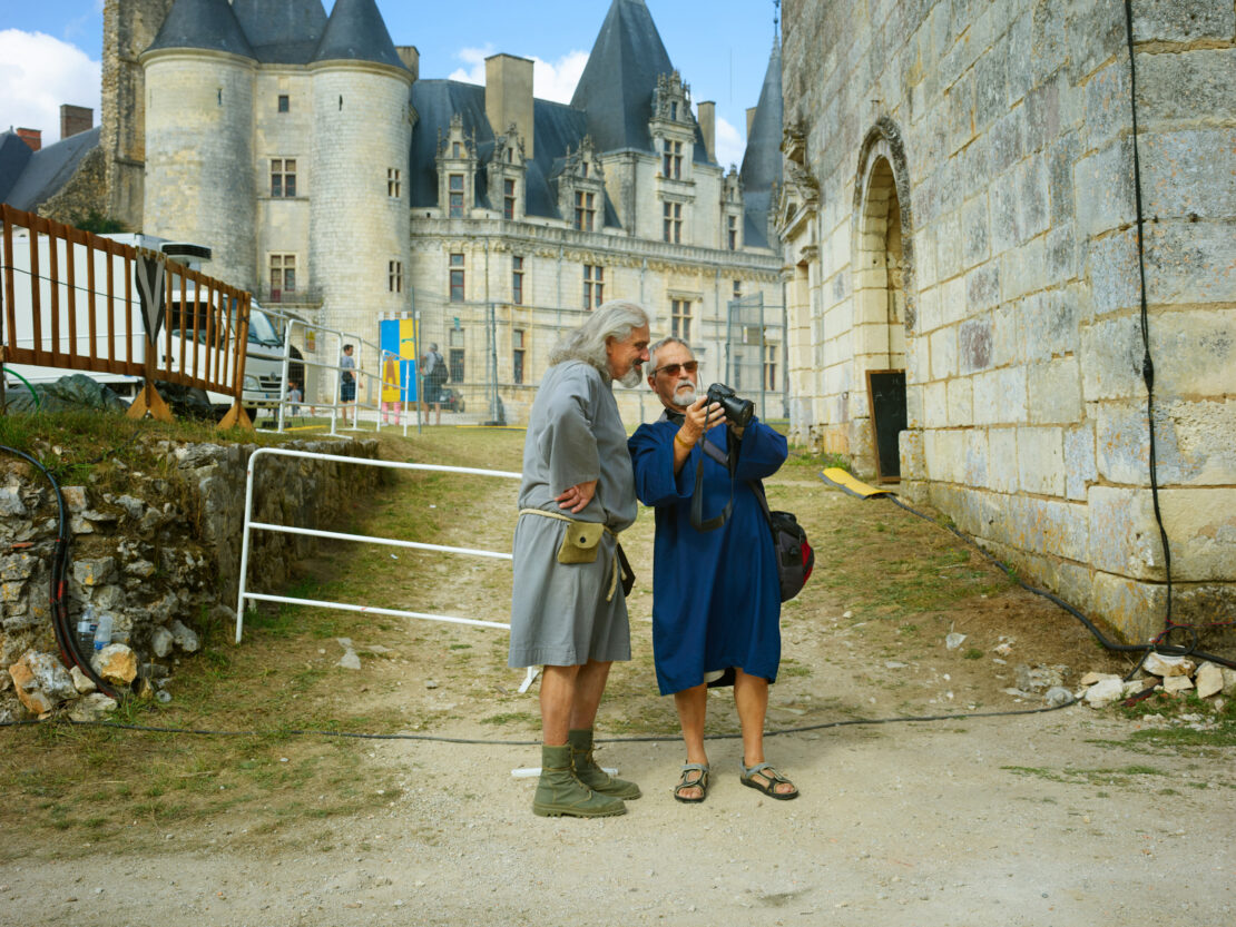 Two men, one photographing a castle, stand on a dirt path in daylight. One is wearing sandals and blue robe and the other period costume.