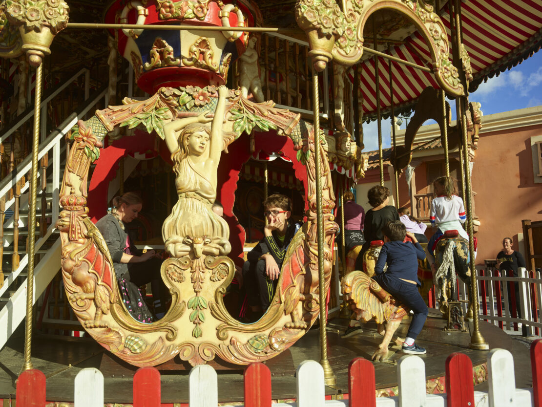 A vintage carousel with detailed carvings is photographed outdoors, featuring both adults and children enjoying the ride on a sunny day.