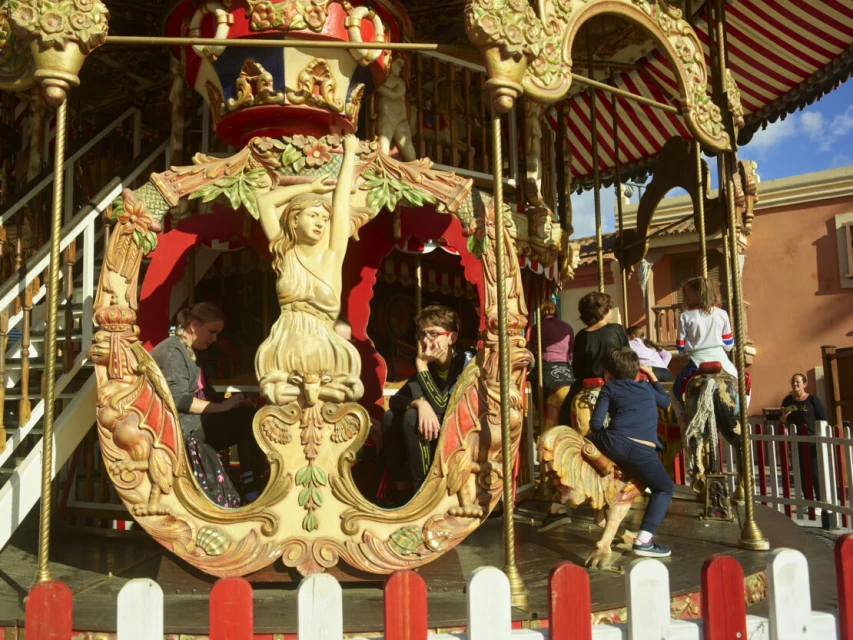 A vintage carousel with detailed carvings is photographed outdoors, featuring both adults and children enjoying the ride on a sunny day.