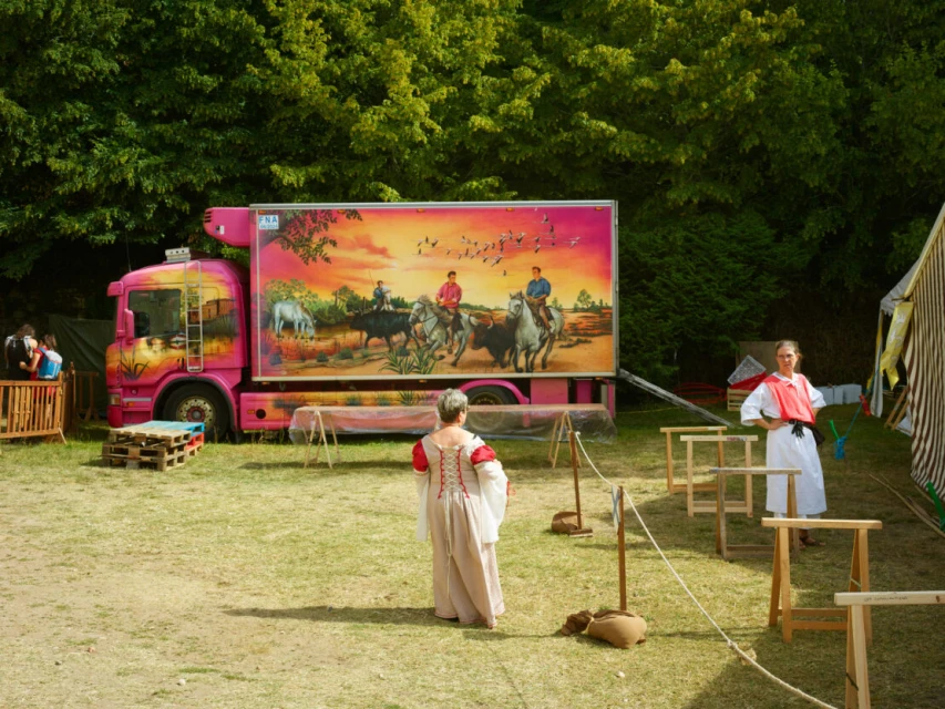 Outdoor scene featuring a pink truck with a horse and rider mural, two women in period costumes, and wooden structures on a grassy field.