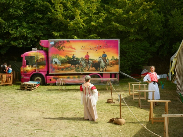 Outdoor scene featuring a pink truck with a horse and rider mural, two women in period costumes, and wooden structures on a grassy field.