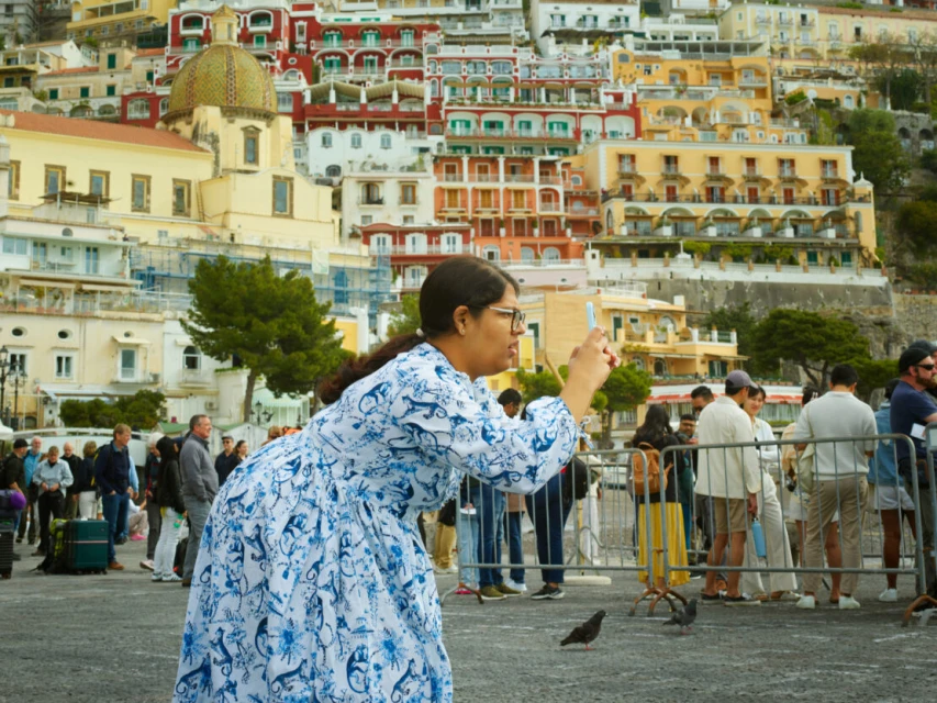 Woman in blue and white dress taking a picture in Positano, Italy, with colorful buildings and people in the background.