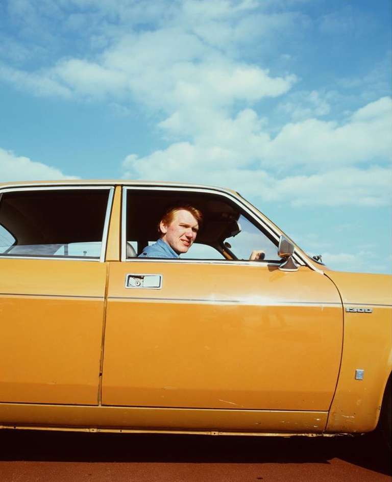 A red-haired man smiles and drives a vintage yellow car with a bright, partly cloudy sky visible through the windows behind him.