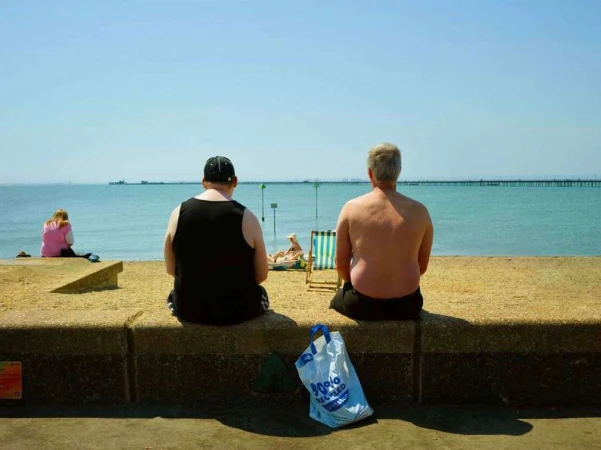 Two men sit seen from behind on a stone seawall overlooking the ocean, with a plastic shopping bag on the ground below.