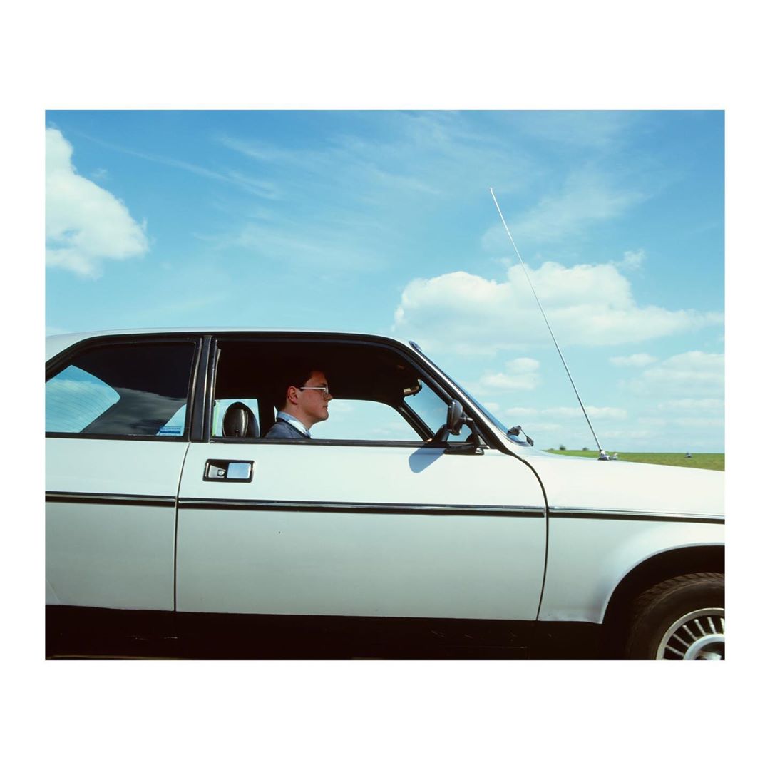 Profile of an adult male wearing glasses and a suit driving a white car on a sunny day with blue sky and light clouds in the background.