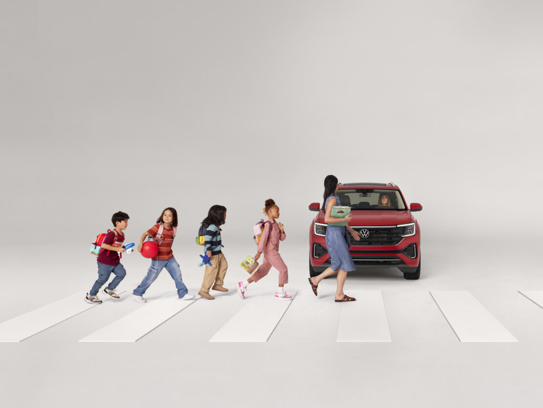 Children cross the street on a crosswalk toward a red Volkswagen SUV, with a woman holding books in this studio advertisement.