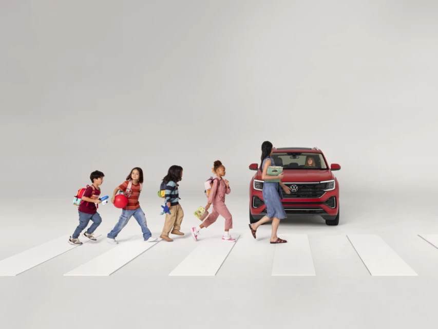 Children cross the street on a crosswalk toward a red Volkswagen SUV, with a woman holding books in this studio advertisement.