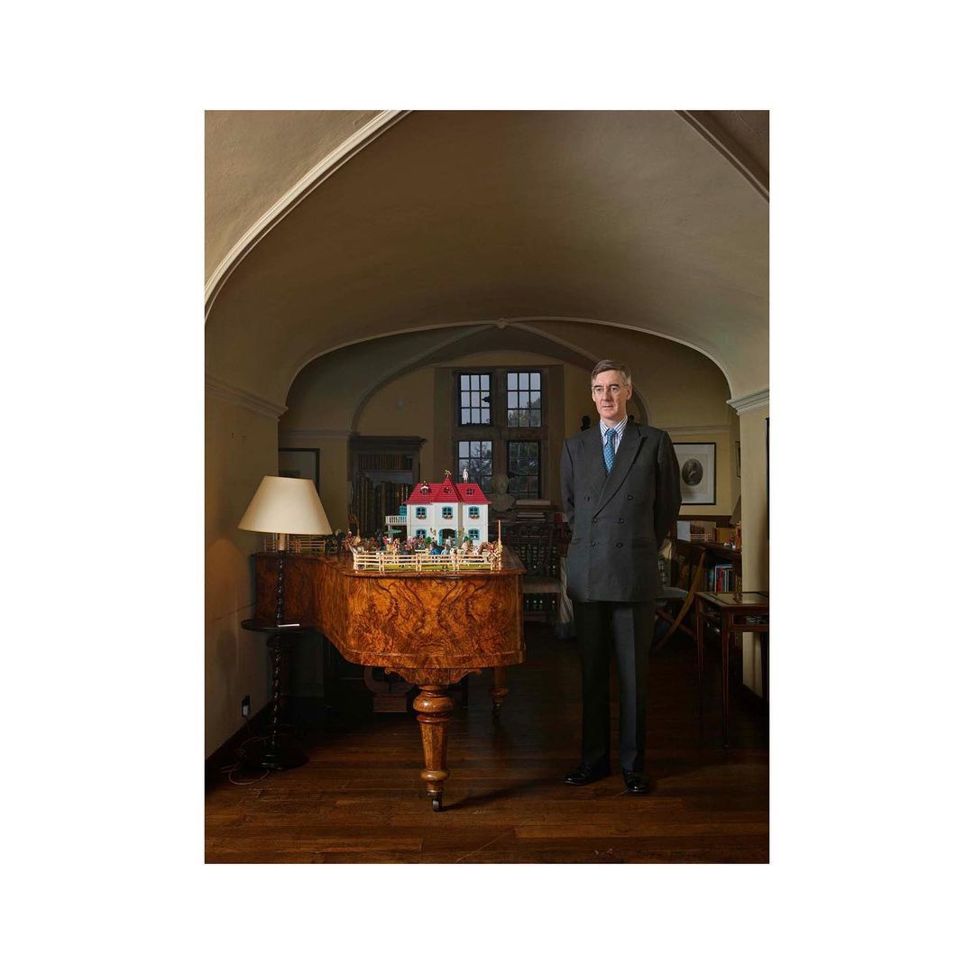 Full-length portrait of an adult man in a suit standing in an indoor space, posed beside a piano and a miniature house display.