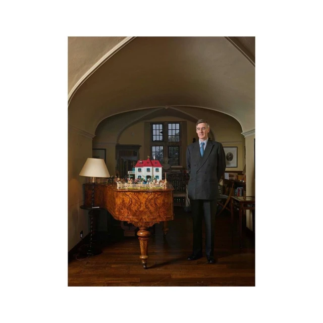 Full-length portrait of an adult man in a suit standing in an indoor space, posed beside a piano and a miniature house display.