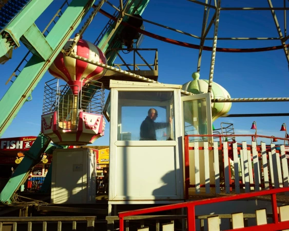 Ferris wheel with colorful cars and an adult man in the operator's booth against a clear blue sky on a sunny day at an amusement park.