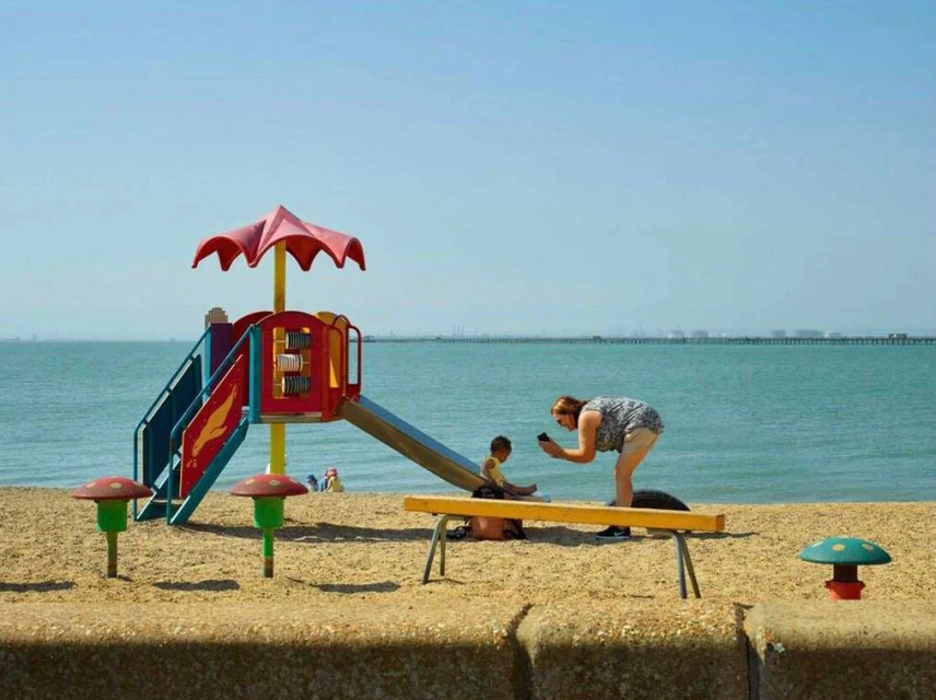 Woman takes a photo of a child on a beach with playground equipment, under clear skies. Beach, ocean, and structure are visible.
