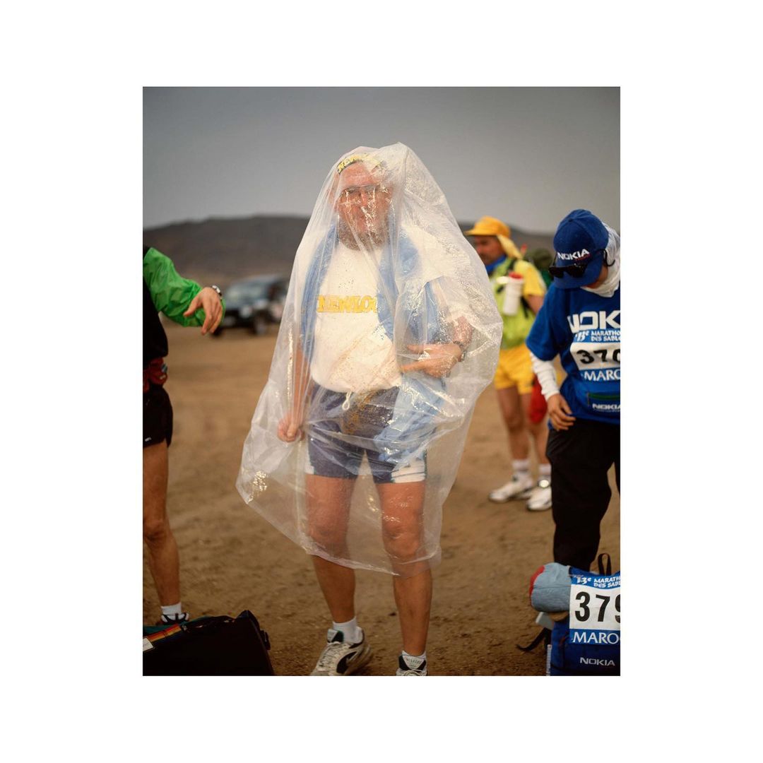 Marathon runner wrapped in a clear plastic sheet, standing with other runners on a dirt field under an overcast sky, wearing athletic gear.