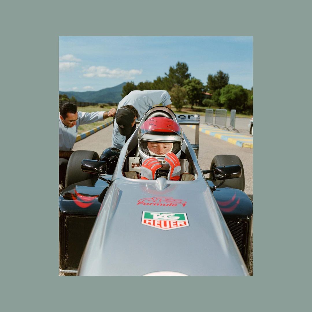 Front-angle shot of a young driver in a racing car, wearing a red helmet and gloves, with two men near the vehicle on an outdoor race track.