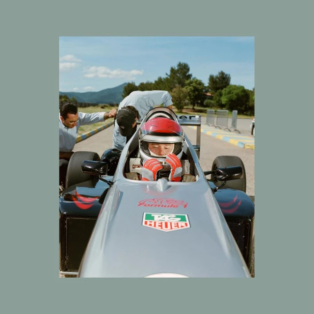 Front-angle shot of a young driver in a racing car, wearing a red helmet and gloves, with two men near the vehicle on an outdoor race track.