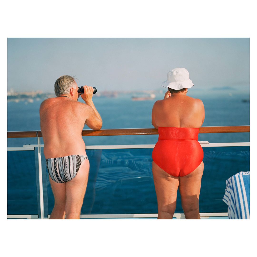 Two people on a cruise ship deck overlooking the ocean. One person uses binoculars, while the other looks out over the water.