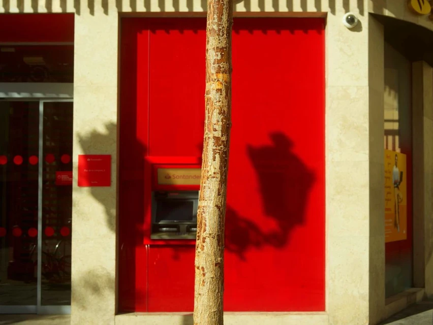 Exterior of a Santander bank with a red wall and ATM, with a tree trunk in the foreground on a sunny day in an urban environment.