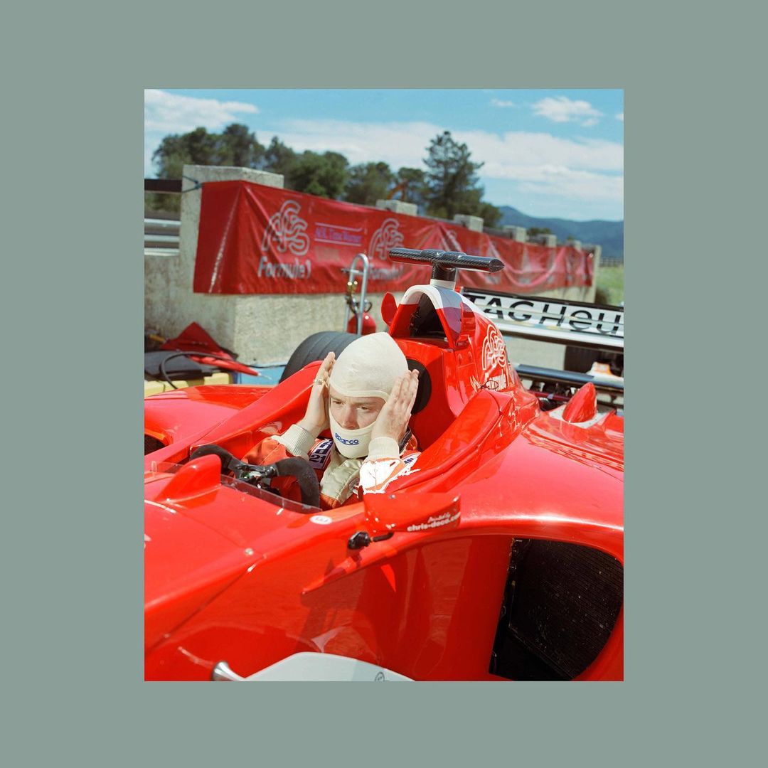 Formula One driver in red race car, hands on head, wearing racing helmet and balaclava on a racetrack, outdoors on a sunny day.
