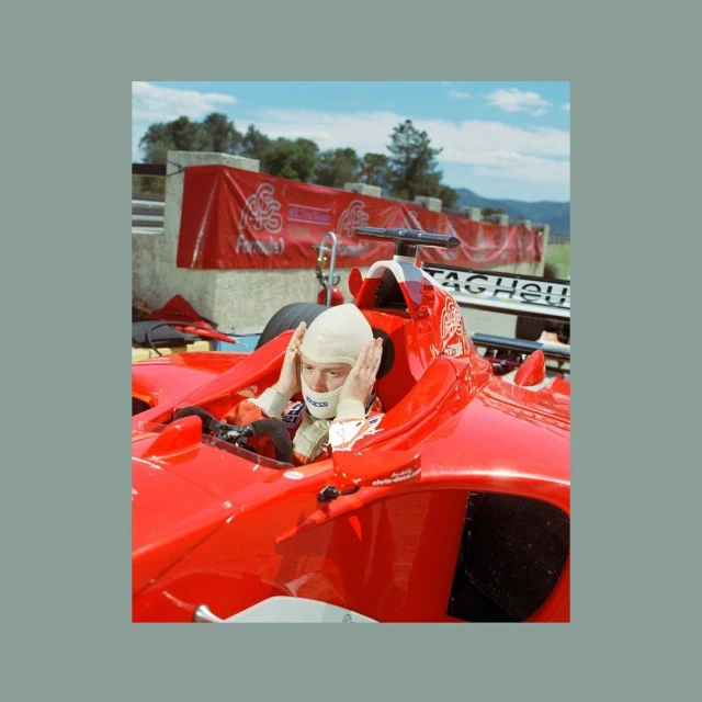 Formula One driver in red race car, hands on head, wearing racing helmet and balaclava on a racetrack, outdoors on a sunny day.