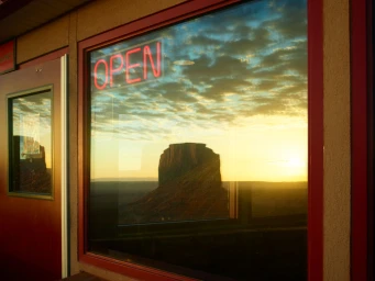 A glowing red neon open sign mounted in a window reflecting a dramatic desert landscape sunset with a flat-topped monolith
