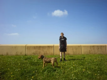 Young boy standing on grassy ground next to a brown dog with a yellow wall and bright blue sky behind them.