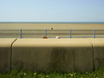 Two people's heads barely visible behind a concrete wall overlooking a wide sandy beach and calm ocean by Dan Burn-forti