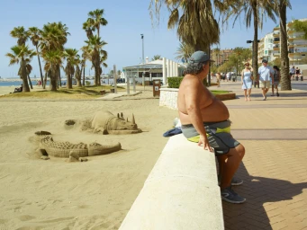 An older man sitting on a low wall next to sand sculptures of a crocodile and a rhinoceros on a sunny beach promenade.