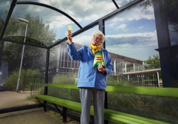 Elderly woman holding a crisp up to inspect it inside a glass bus shelter holding a Walkers crisps packet