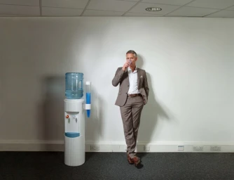 Man in a brown suit drinking from a plastic cup next to a water cooler in an office setting