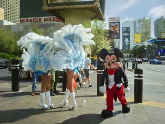 A person in a cartoon mouse costume walks past showgirls wearing large feather headdresses on a sunny Las Vegas street.