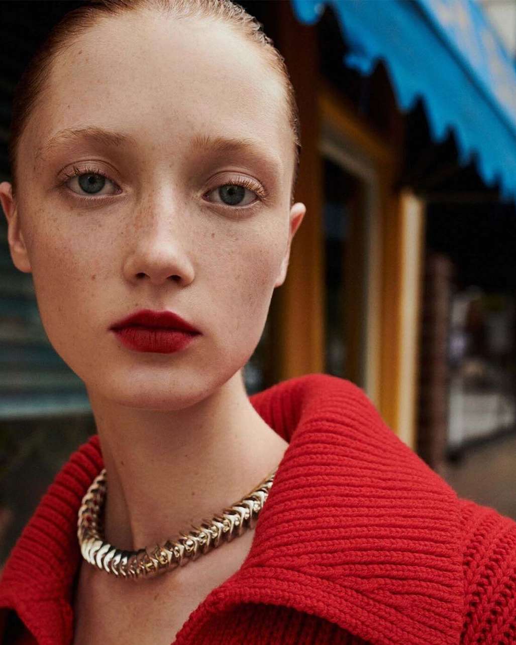 Close-up portrait of a young woman with red hair wearing a red top and gold necklace, posing in front of an outdoor storefront.