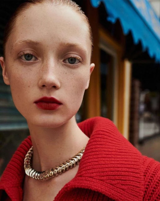 Close-up portrait of a young woman with red hair wearing a red top and gold necklace, posing in front of an outdoor storefront.