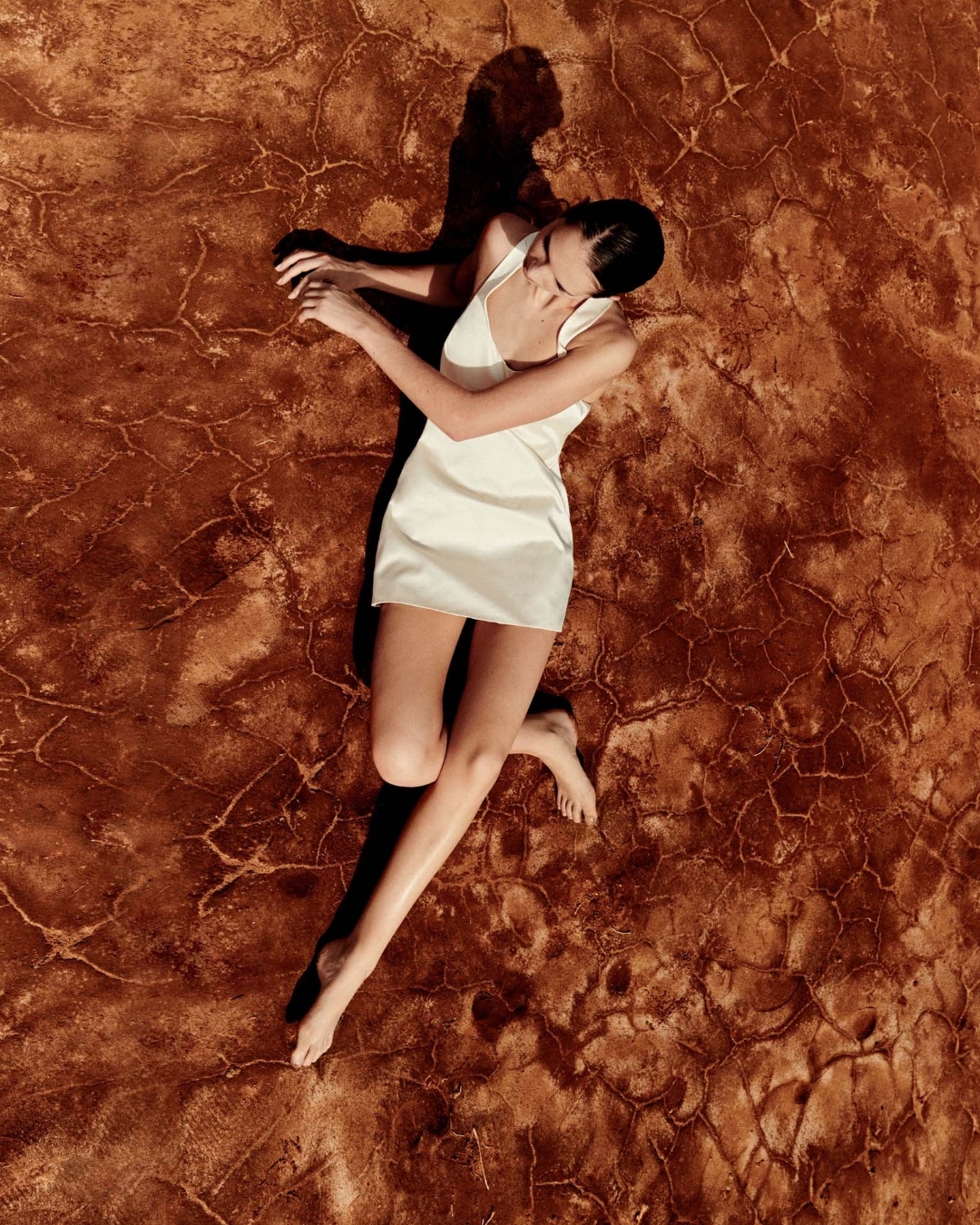Aerial view of a young adult woman lying on a cracked, textured, earth-toned surface, wearing a simple white dress in soft sunlight.