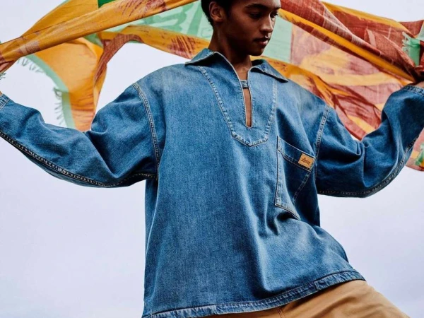 Young adult in a denim collared shirt posing with a colorful patterned scarf against a gradient sky, fashion forward attire.