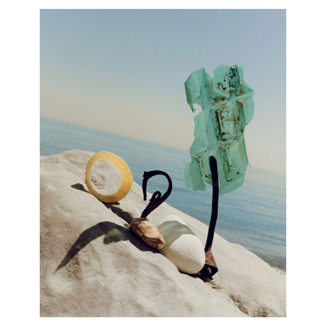 Still life of unique objects including a ring, rock, metal fixture, and glass art against ocean background on a stone surface.