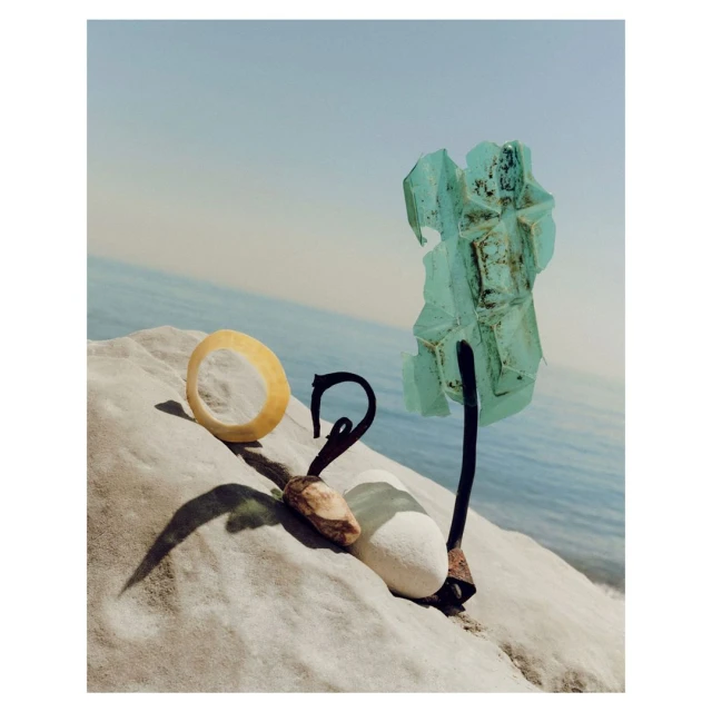 Still life of unique objects including a ring, rock, metal fixture, and glass art against ocean background on a stone surface.