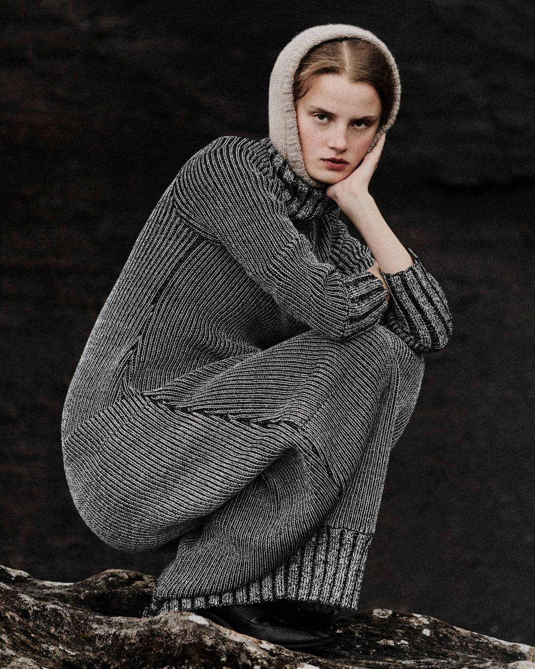 Fashionable woman in grey knitwear and a hood, sits on a rock against a dark background, showcasing fashion, style, and monochrome.
