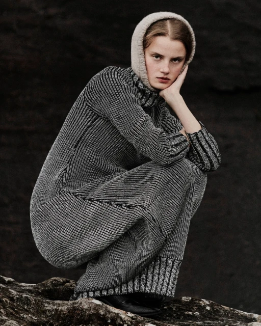 Fashionable woman in grey knitwear and a hood, sits on a rock against a dark background, showcasing fashion, style, and monochrome.