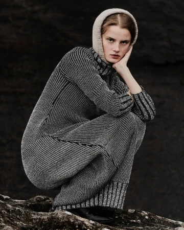 Fashionable woman in grey knitwear and a hood, sits on a rock against a dark background, showcasing fashion, style, and monochrome.