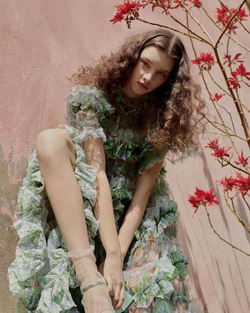 Woman with long curly brown hair wearing a ruffled tulle dress posed outdoors near a pink wall and red flowers.